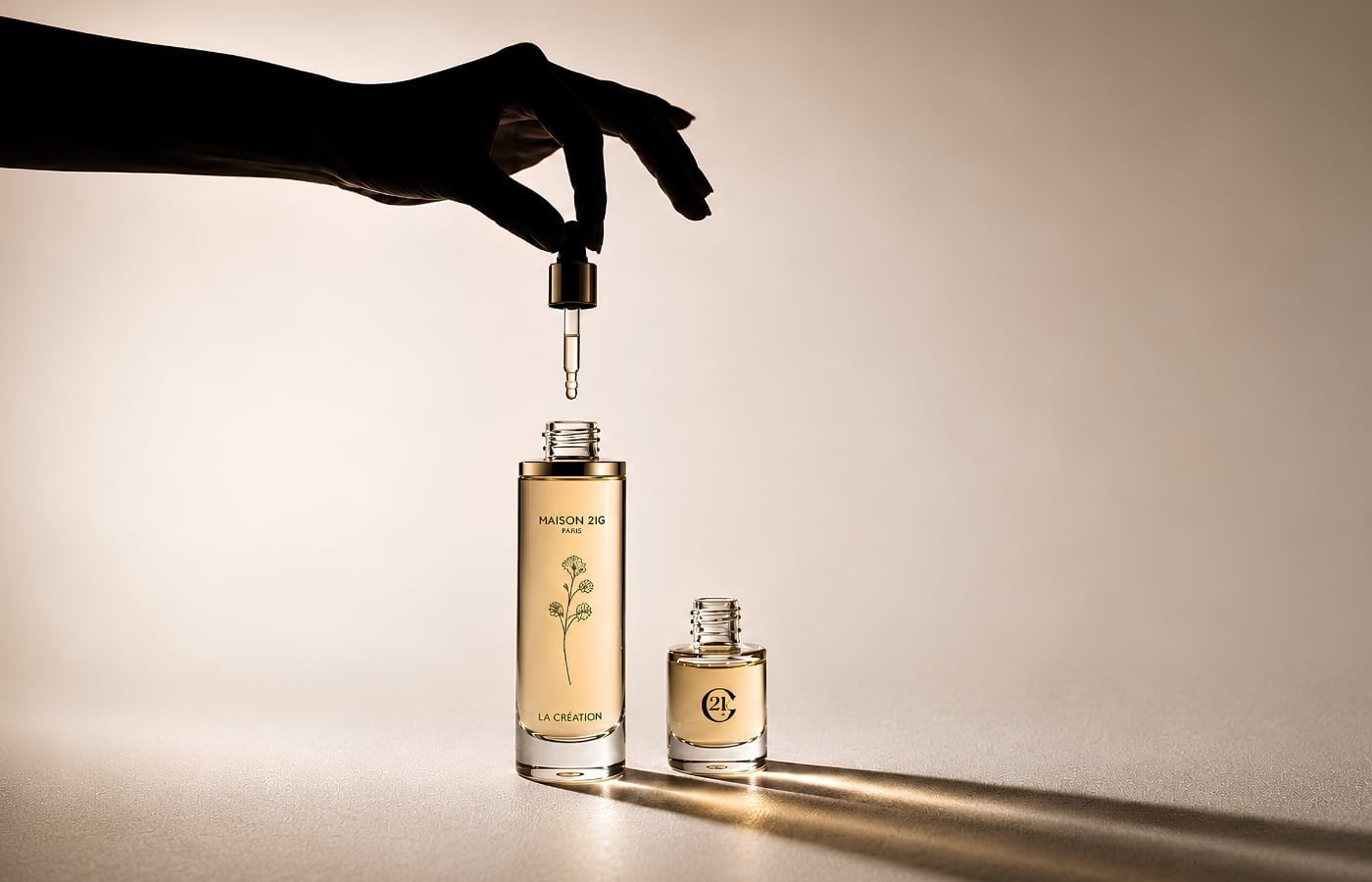 A silhouetted hand lets a single drop fall from a gold dropper into a Maison 21G Paris La Création bottle, a Scent Magnifier vial beside it, in warm champagne backlight.