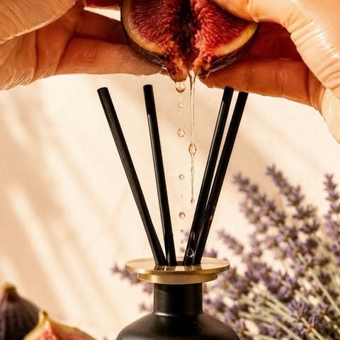 A hand squeezes a ripe fig above a black reed diffuser, with Lavender beside it.