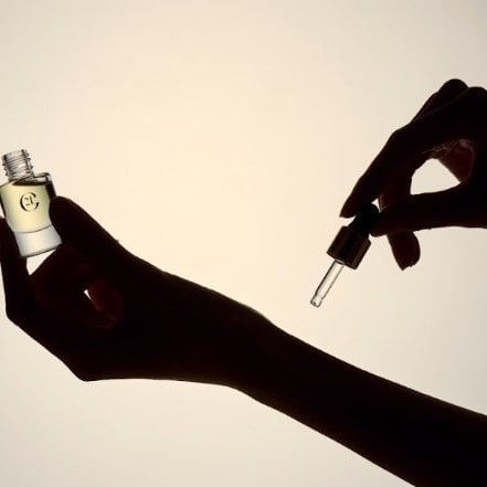 Two silhouetted hands against warm light: one holds the open Scent Magnifier vial, the other its dropper.