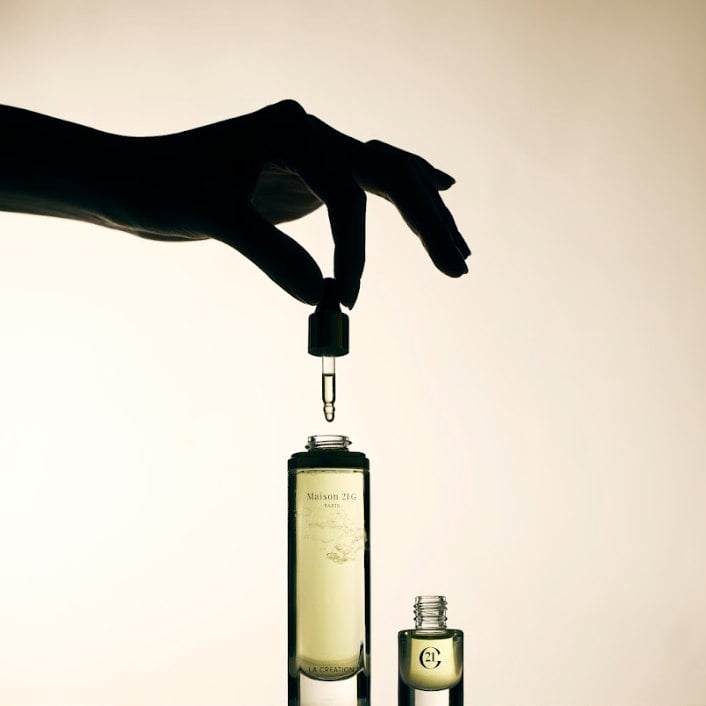 A silhouetted hand releases a single drop from the Scent Magnifier dropper into an open bottle of La Création Eau de Parfum.